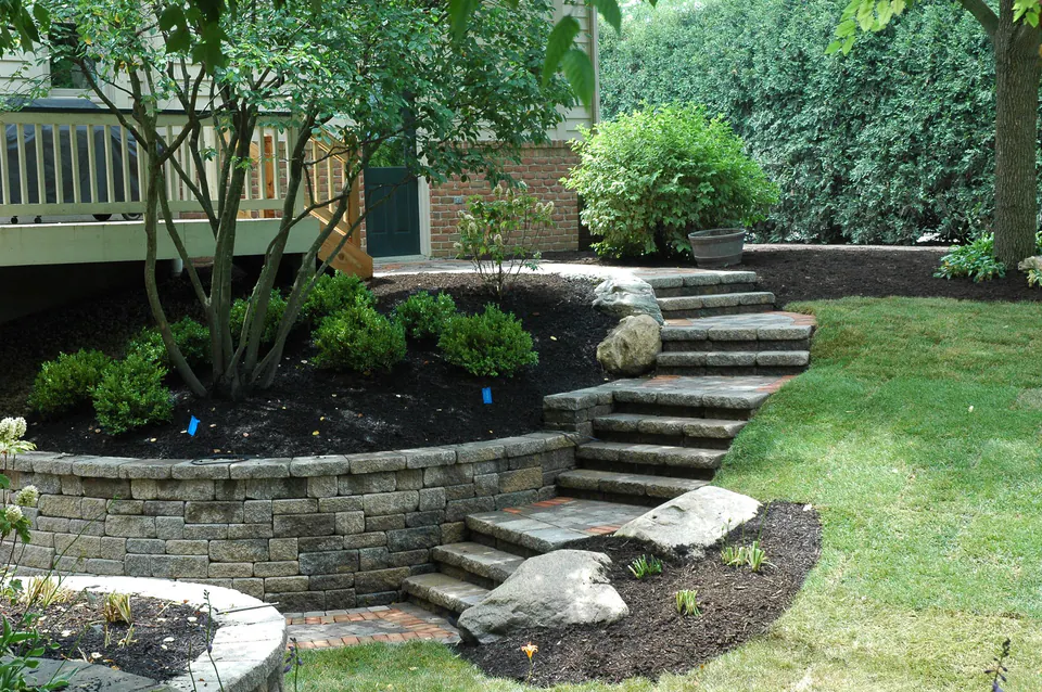 Segmental block retaining wall with stone steps curving around a planting bed near a deck